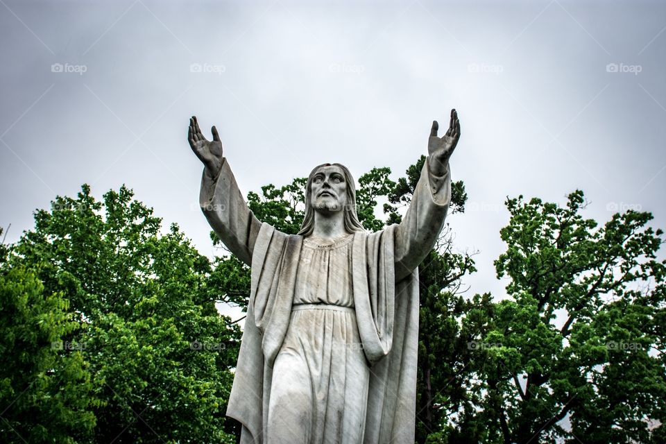 Sculpture/statue of Jesus outside with his arms raised up surrounded by greenery on an overcast day.