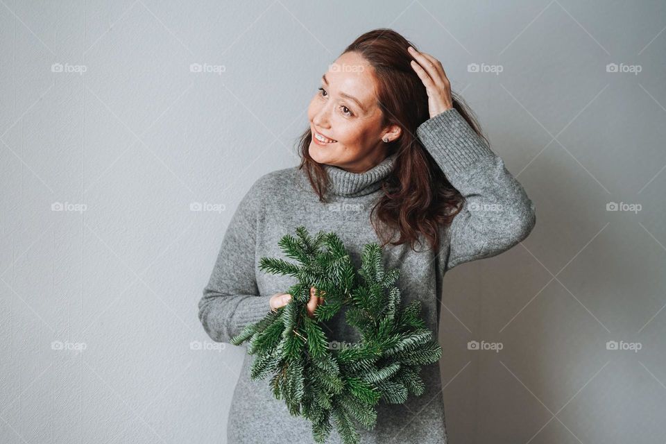 Adult beautiful smiling woman forty years with brunette curly hair in warm grey knitted dress with diy fir christmas wreath in hand on grey background