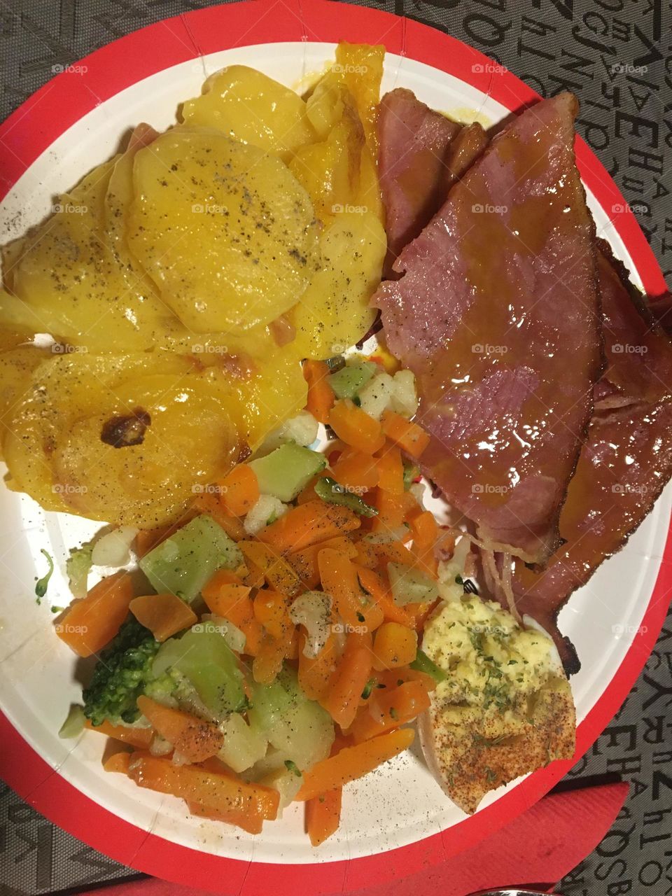 Delicious plated homemade supper consisting of honey mustard glazed sliced ham, cheesy scalloped potatoes, and boiled mixed vegetables 