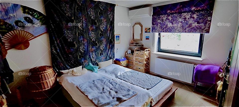 This is the bedroom in the lovely bungalow in “Bavaria”, Germany. A big ornamented tapestry hangs from the wall behind the bed. Matching curtains, a mirror, a wicker rattan chest with drawers complete the interior design. 2023. Hypnotic Productions