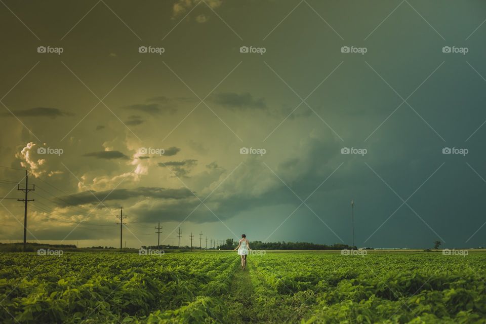 A women in a white dress walks through a Manitoba prairie field. The sun is setting over a cloudy sky, which creates a dramatic landscape.