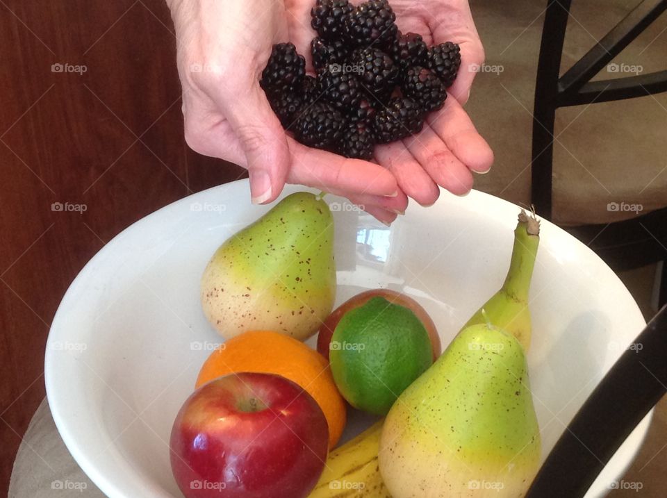 Holding in hand blackberries near a bowl of colorful fruit.