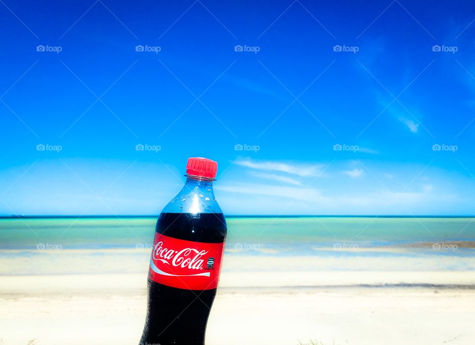 A vivid tropical image of an unopened bottle of Coca Cola  with its contents lining up with the ocean horizon. 