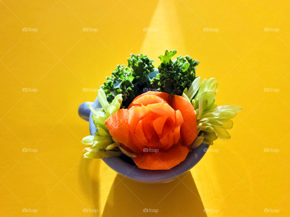 My amateur attempt at an orange carrot rose nestled together with dark green curly kale & pale green flower petals in a tiny blue teacup on a bright yellow background. The early evening sunlight produced a triangular light beam & shadow.