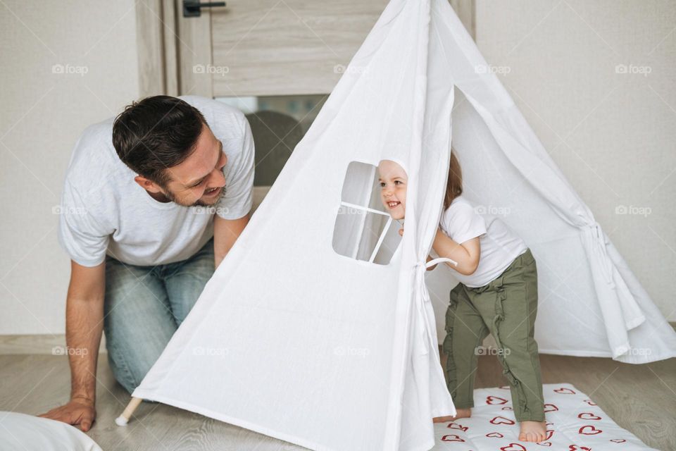 Happy father young man and baby girl little daughter having fun playing in wigwam in children room at home