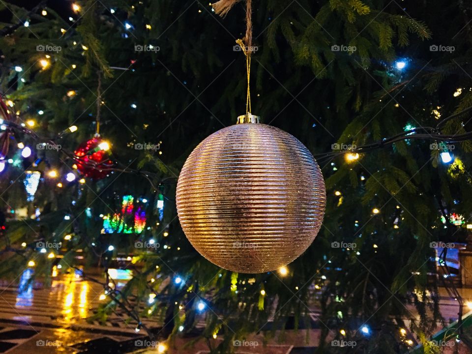 Giant golden Christmas globe hanging on the Christmas tree outside with lights in the background 