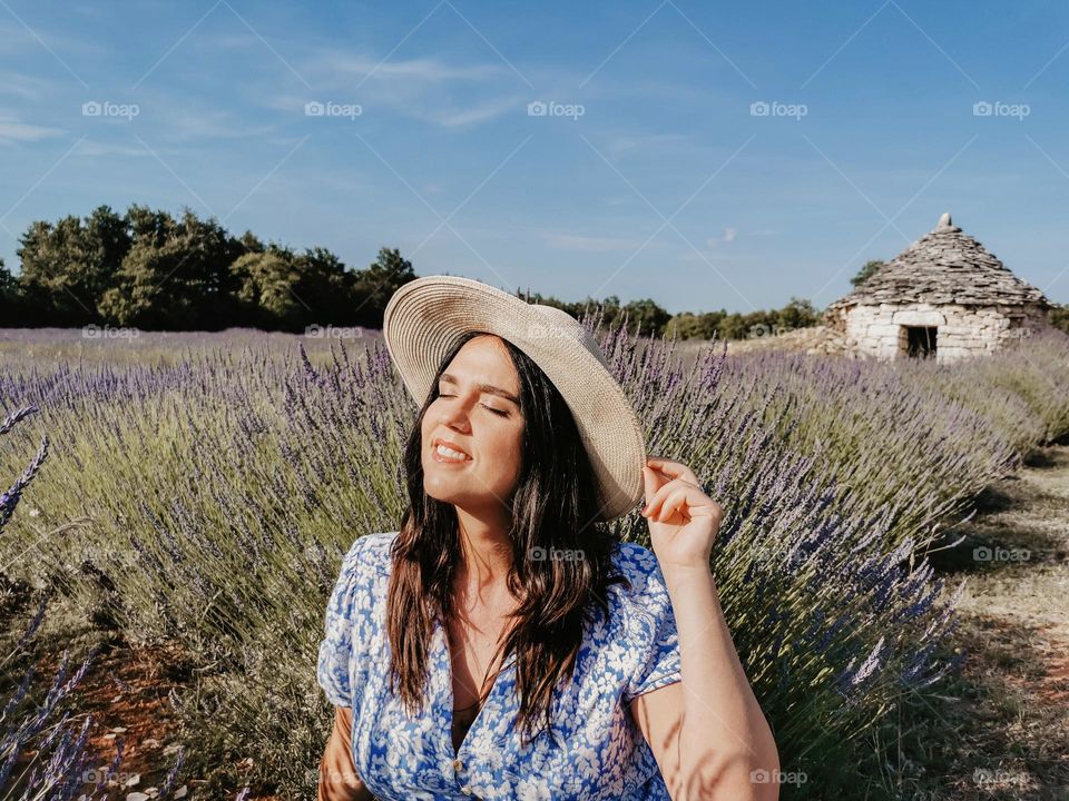 Portrait of happy young woman wearing dress and sun hat sitting in a lavender field