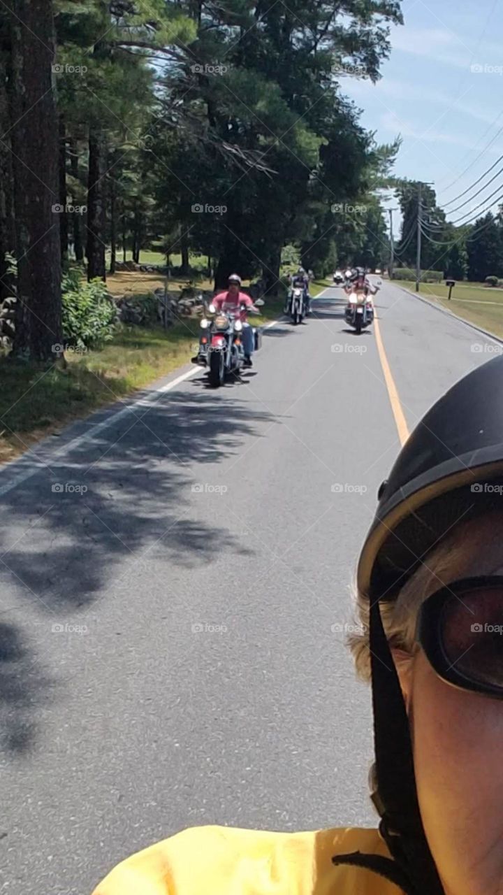 Roadway behind rider on motorcycle run in summertime. Passenger wearing helmet, sunglasses, motorcycles in road behind lead bike. Motorcycles riding behind biker taking selfie.