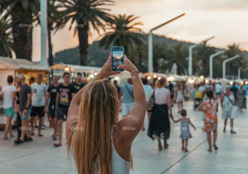 Rear view of woman standing in crowded street in city, taking photos with mobile phone