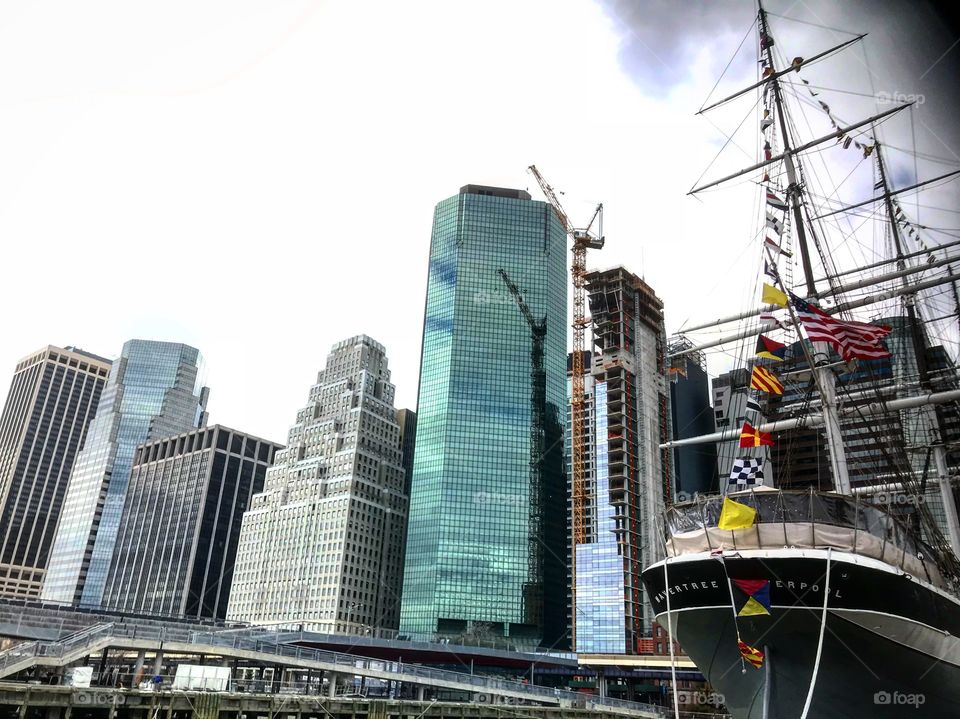 Photo of Wall Street and the financial district of NYC  taken from South Street Seaport on a cloudy day. A large ship is seen in the right foreground. 