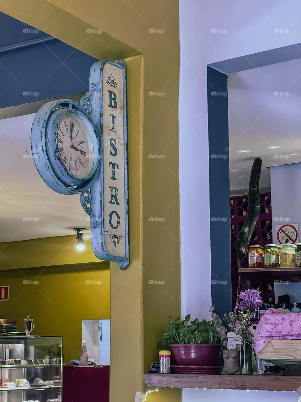 Um relógio de parede estilo vintage com detalhes rústicos dentro de um aconchegante café bistrô. As paredes amarelas e brancas fornecem um pano de fundo aconchegante para a atmosfera convidativa, realçada por plantas e elementos de decoração.