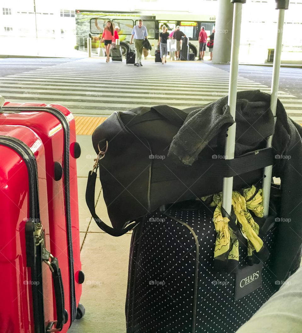 Perspective shot suitcases at arrivals outdoor at airport on curb at crosswalk, travellers in background walking across crosswalk toward the suitcases