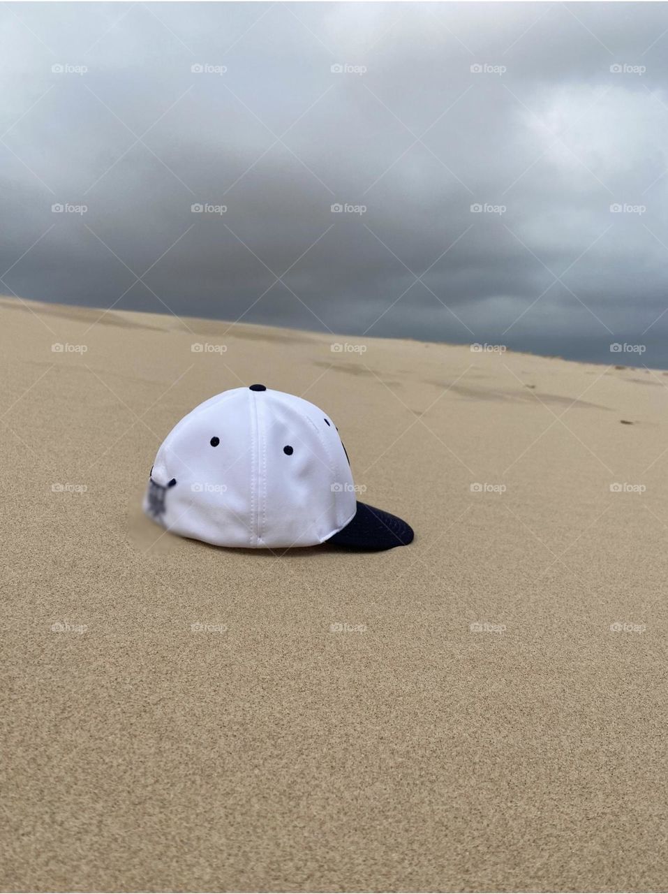 A white and navy blue baseball cap lying on a sandy surface, with a cloudy sky in the background. The setting is a beach with dunes.