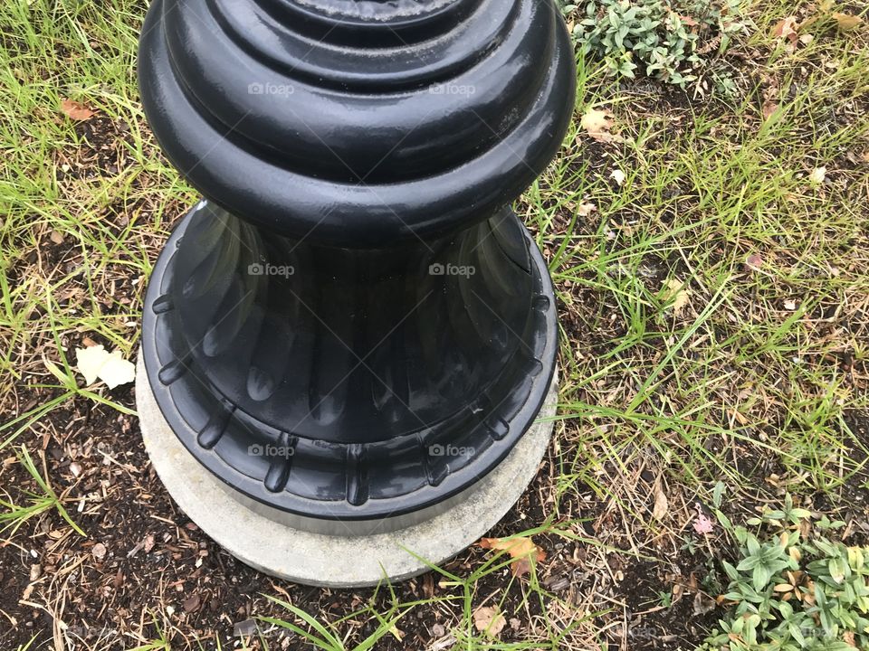 The bottom of a parking lot, street light that is painted black on s cement slab. With grass and dirt surrounding the base it makes for a stark contrast. 