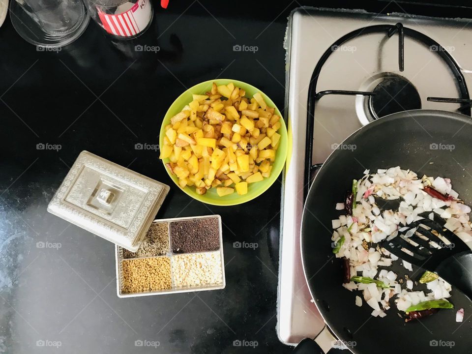 A pan with frying onions and plate full of chopped potato’s and four compartments spice’s box on kitchen counter.