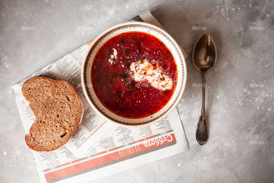 Plate of Ukrainian borsch. Nearby are two sliced ​​pieces of bread and a spoon. A plate is on the newspaper. Photo taken on a white concrete background.Red borsch, add sour cream and greens.