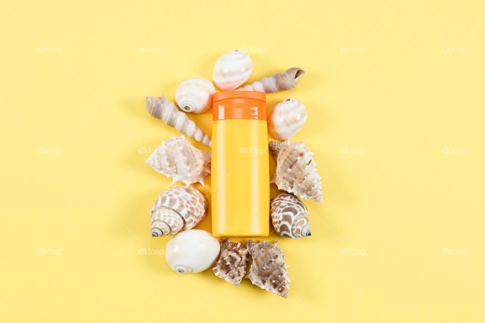 One small tube of suntan cream with shells around it lies in the middle on a yellow background with copy space on the sides, flat lay, close up.