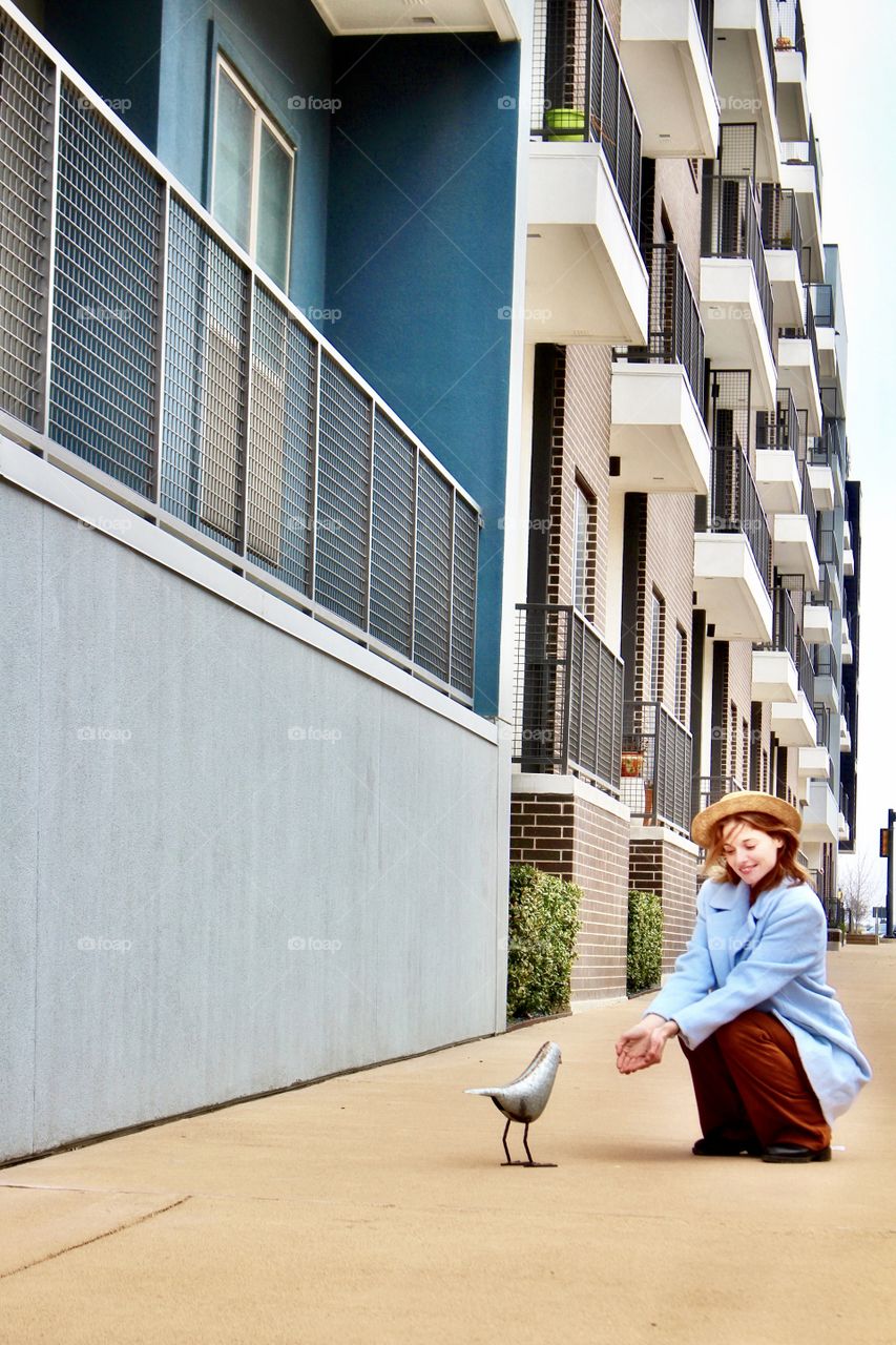 A young millennial woman in the city offering food to a bird and surrounded by geometric architectural buildings in a urban setting!