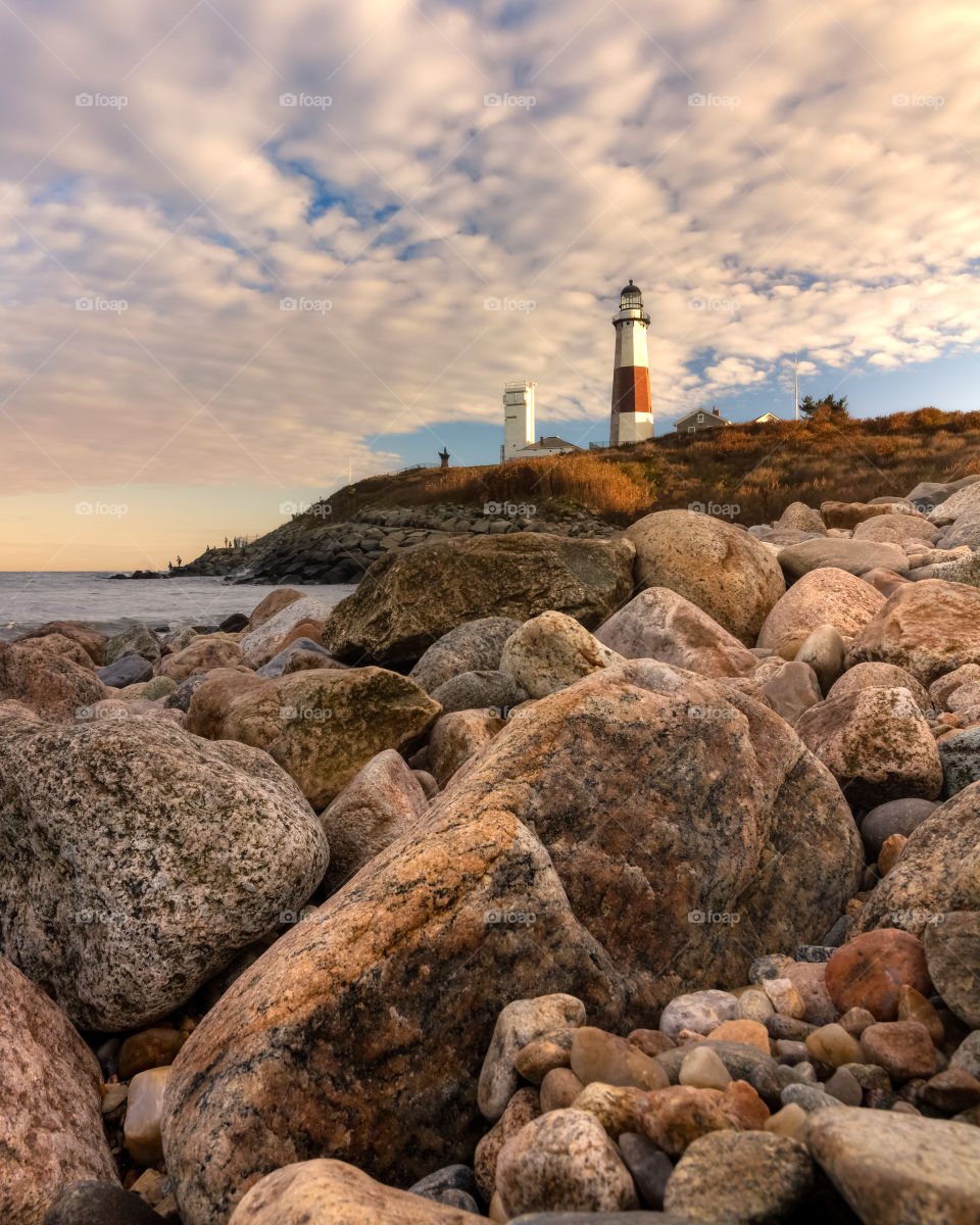 Warm golden hour tones light up the side of a lighthouse and rocky coastline. Montauk Point, Long Island New York.