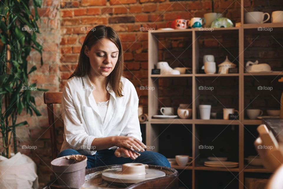 Young attractive woman in white shirt ceramic artist working on potter's wheel in studio. Handmade work student, freelance small business, hobbies and crafting