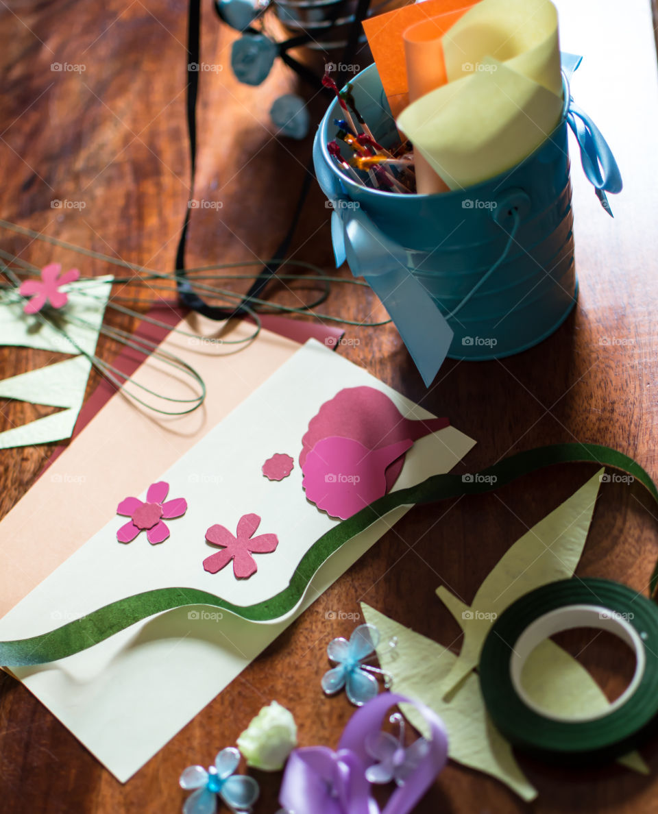 Arts and crafts supplies in bucket on wooden table with supplies spread out to make greeting cards and paper flowers on desktop 