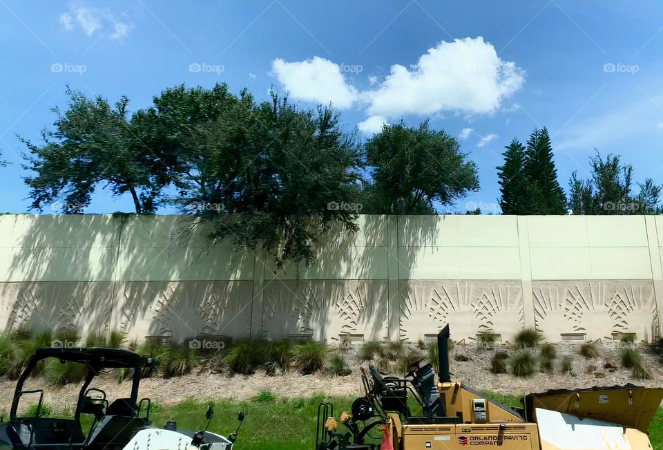 Commuting: vehicles bogies motorized cart and yellow machinery on the highway by a wall with designs and trees peaking out with a beautiful blue sky and clouds.