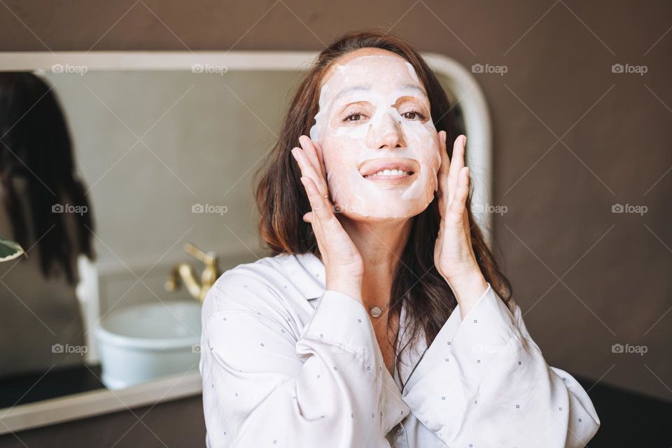 Adult brunette woman with sheet mask on her face in the bathroom at home