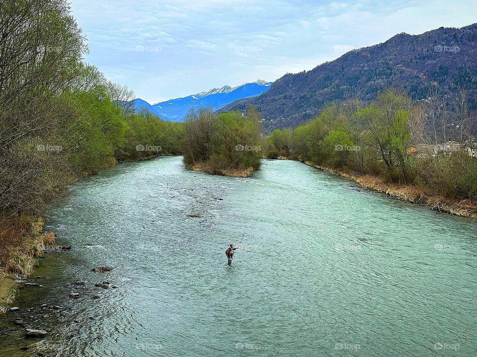 Spring river, which has become shallow. There are green trees along the banks. In the middle of the river stands a fisherman with a fishing rod and catches fish. Mountain peaks are visible in the background.