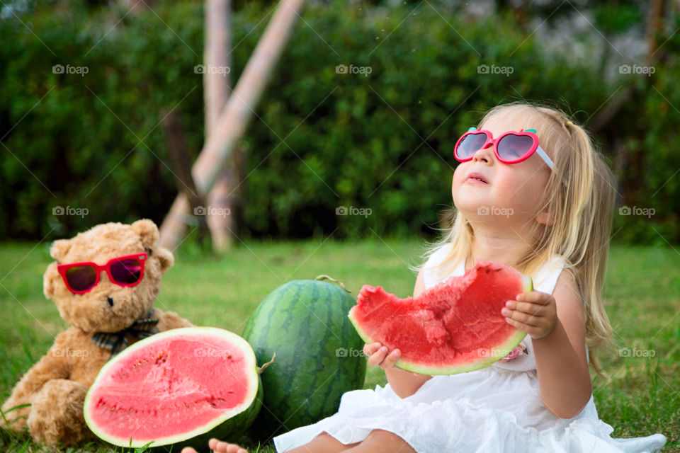 Cute little girl with blonde hair in sunglasses sitting in the park with her teddy bear and eating fresh watermelon 