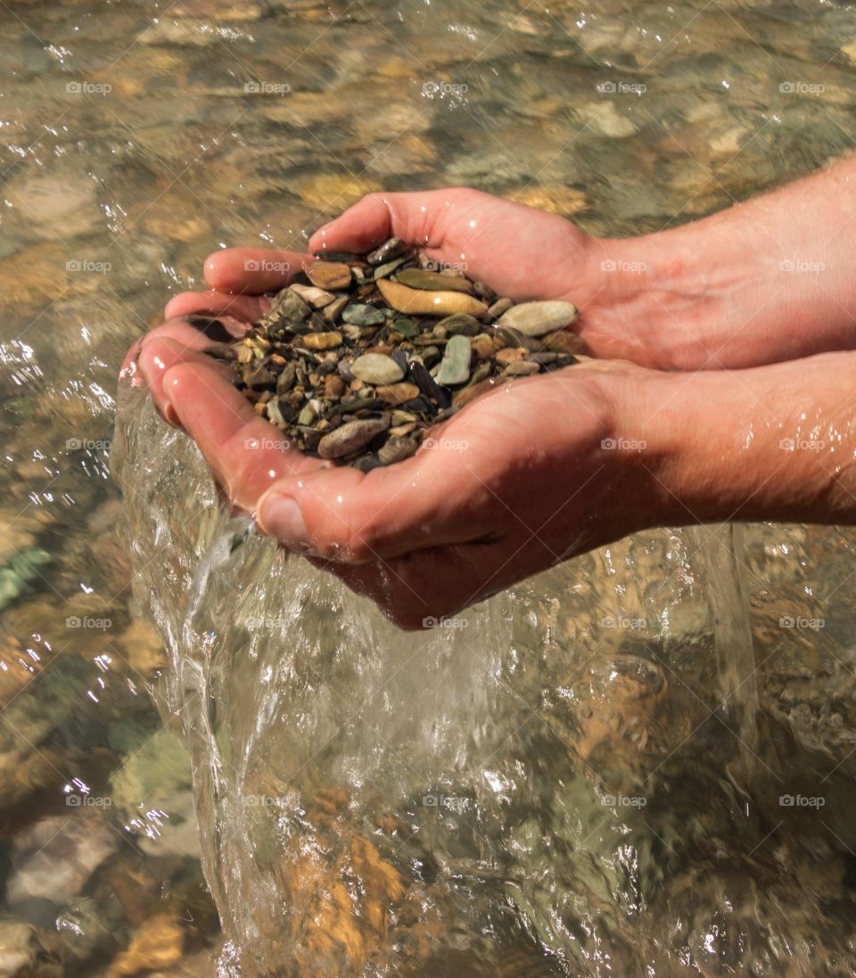 A pair of hands scooping out rocks from the bottom of a shallow creek, with water pouring out of the hands.
