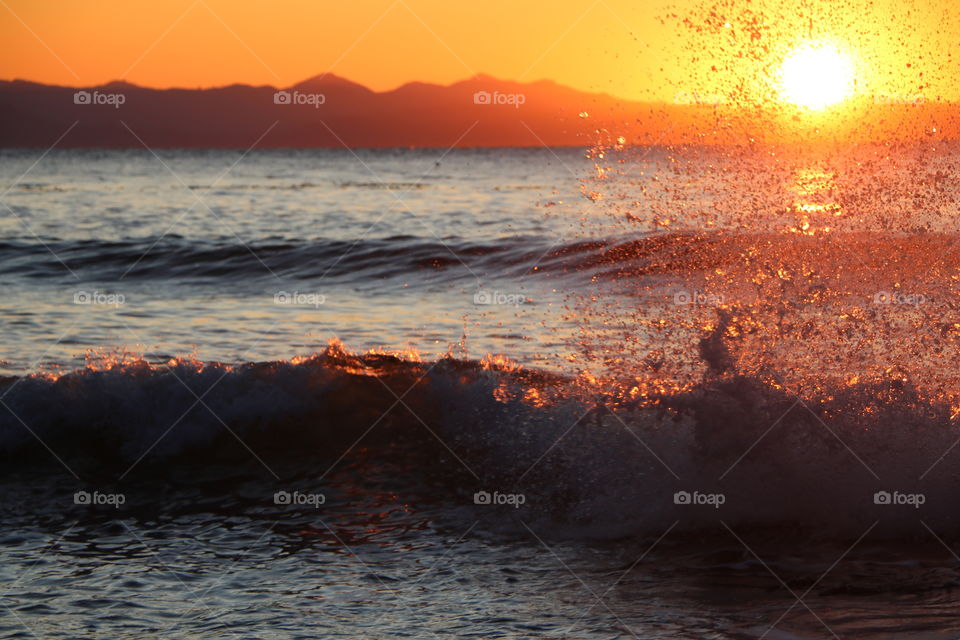 Waves crashing near the shore , water droplets splashing and shining on the golden light from the setting sun