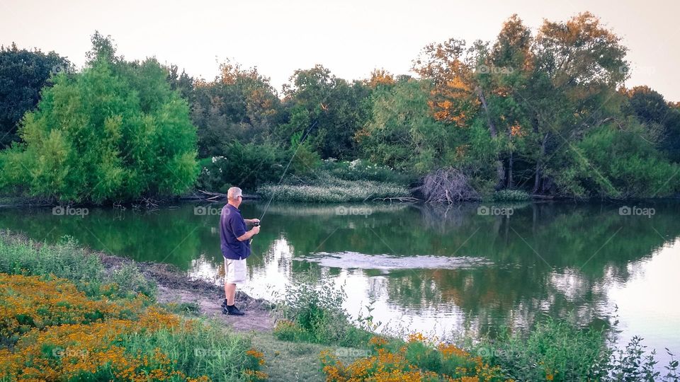 A man fishing in a countryside pond in early fall with trees & flowers and ripples on the water after casting his line