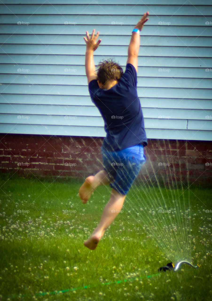 Young boy jumping through backyard sprinkler in summer.