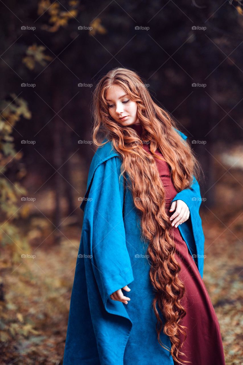 Beautiful girl with long red hair in blue coat. Woman staying in the forest