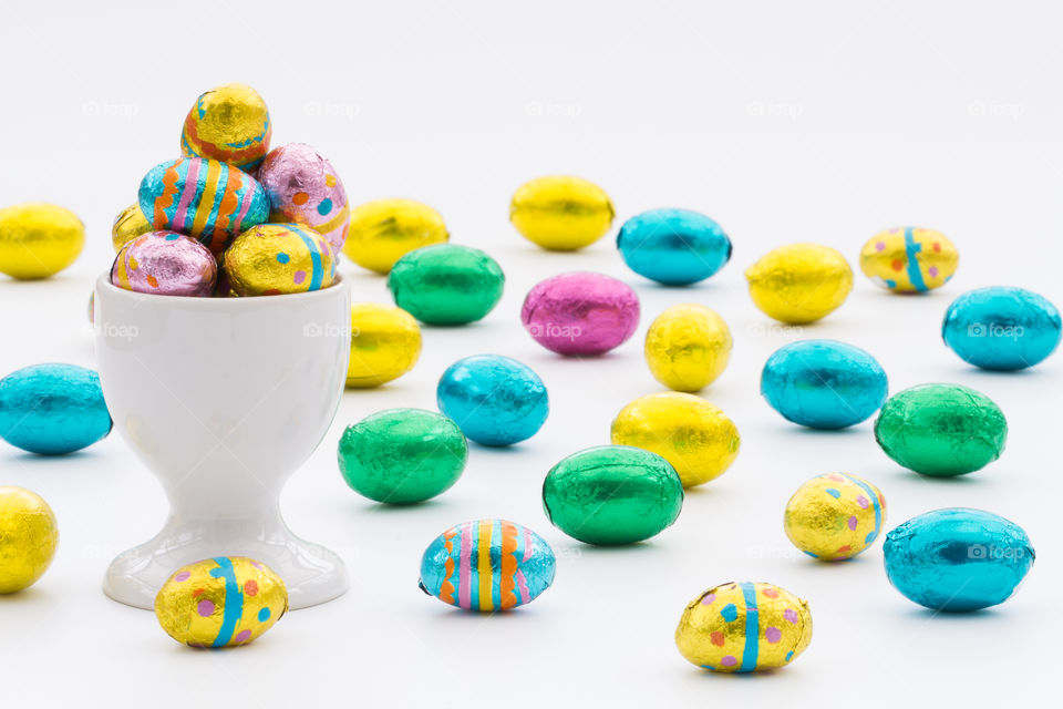 An egg cup full of colourful, chocolate Easter eggs on an isolated white background with lots of Easter eggs scattered around.