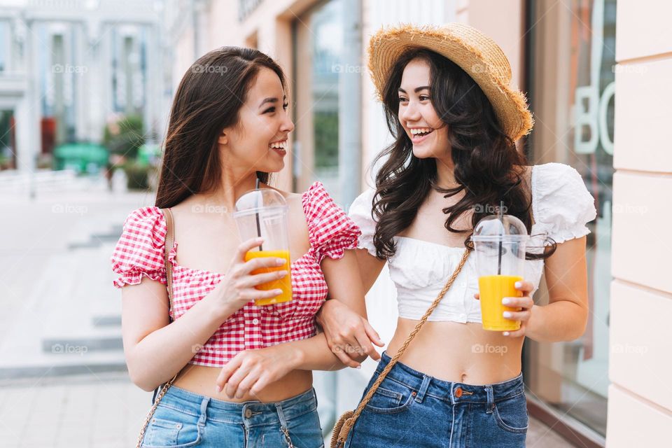 Happy smiling beautiful brunette young women friends in summer clothes with juice in hands walking on the summer city street