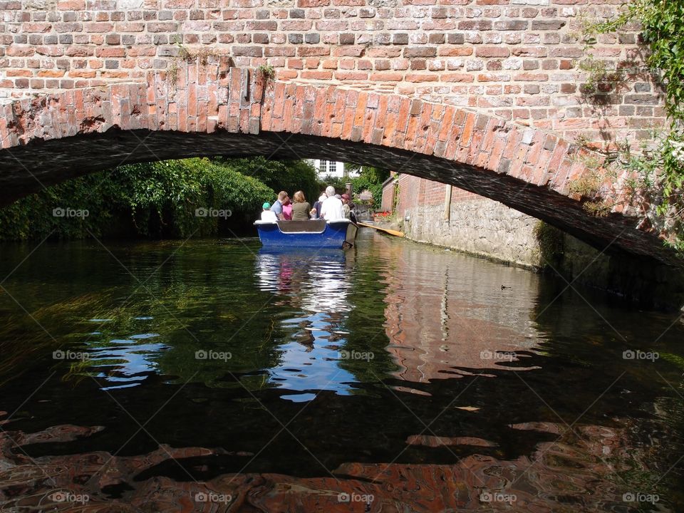 A small tour boat full of people floats down a river in England on a sunny summer day with a ornamental brick bridge in the foreground. 
