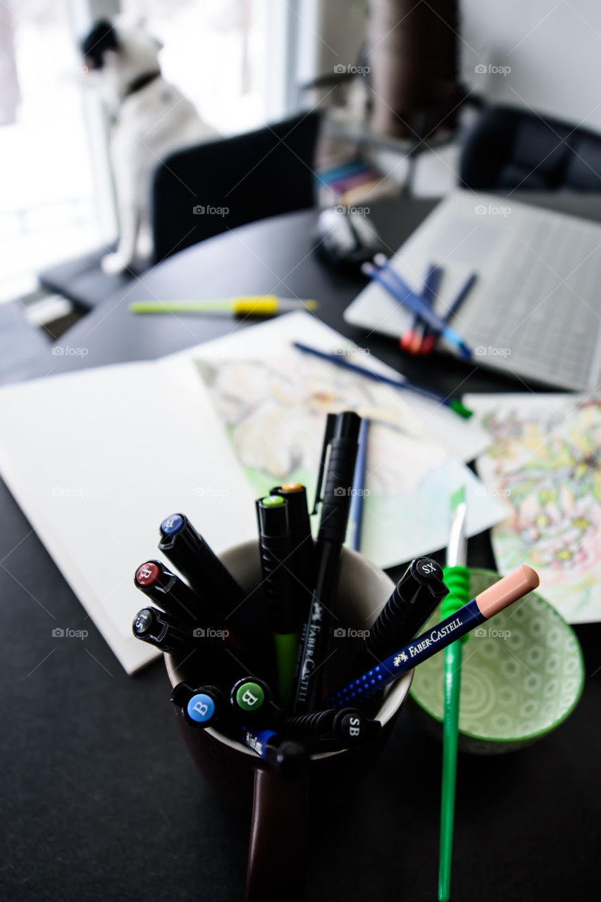 Faber-Castell Art Grip Aquarelle pencils and PITT Artist pens in coffee cup on desk with sketches, sketchbooks, paint brushes, water cup, laptop near window creative workspace and lifestyle conceptual photography background 