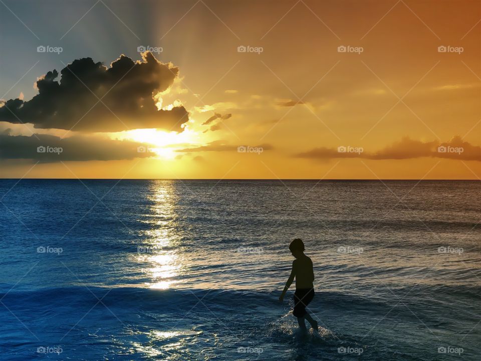 The golden setting sun lights a boys walk through the ocean.