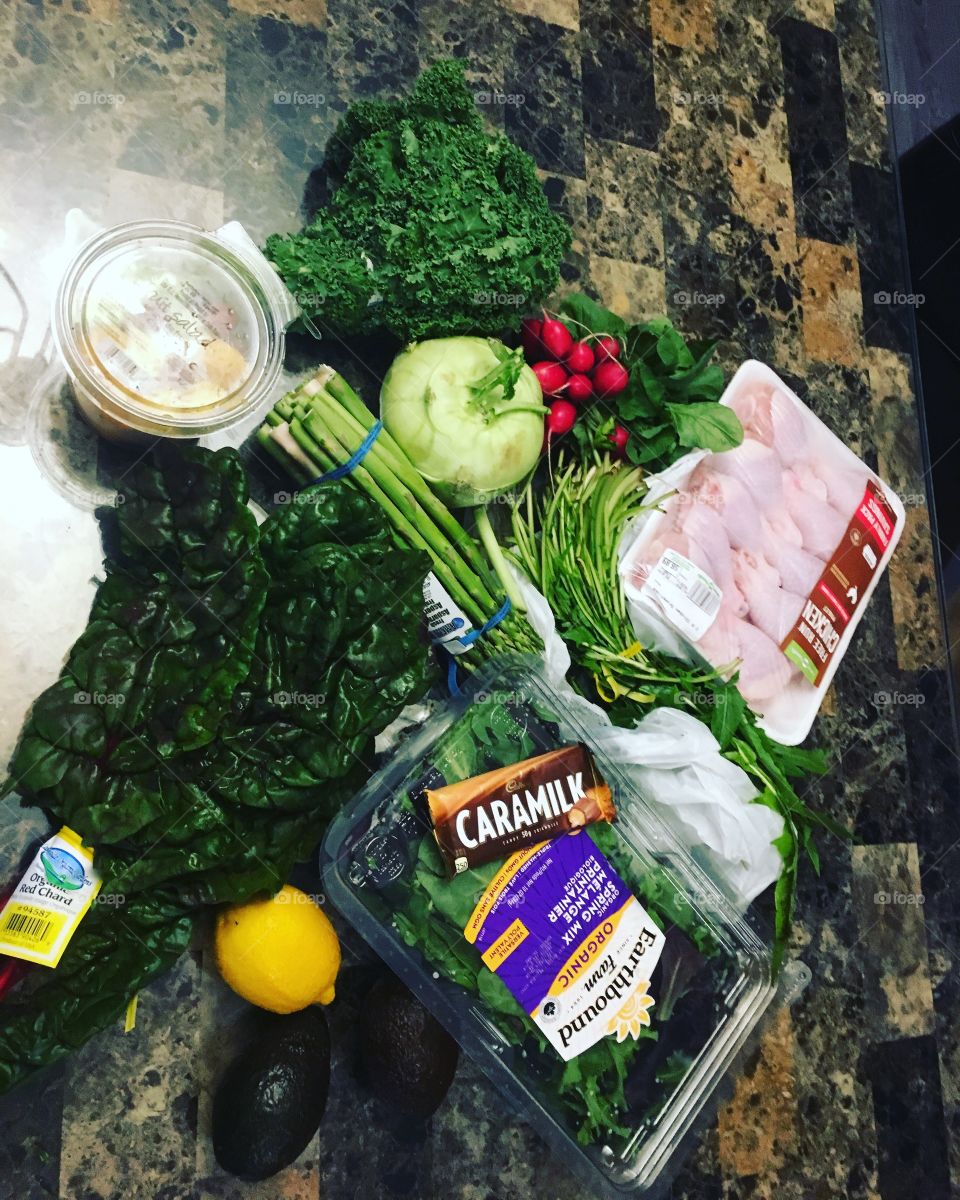 A pile of many groceries, mainly vegetables is spread on a counter. On top of a spring mix is a Caramilk chocolate bar