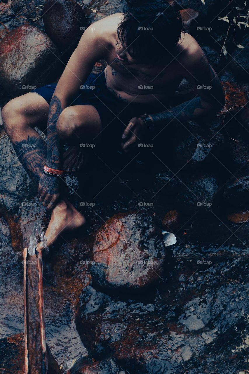 Under a bamboo shower, a tattooed man sits on a mossy rock holding his right leg, both hands and feet covered in tattoos.