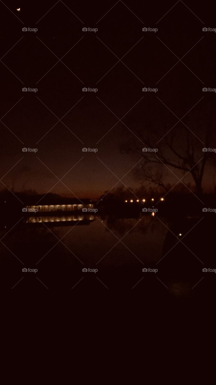 Night photo 2.2 position zoom shore lights office area reflections of trees on Calm Lake Waters. Backlit still visible, reflection of Backlit on Lake Water