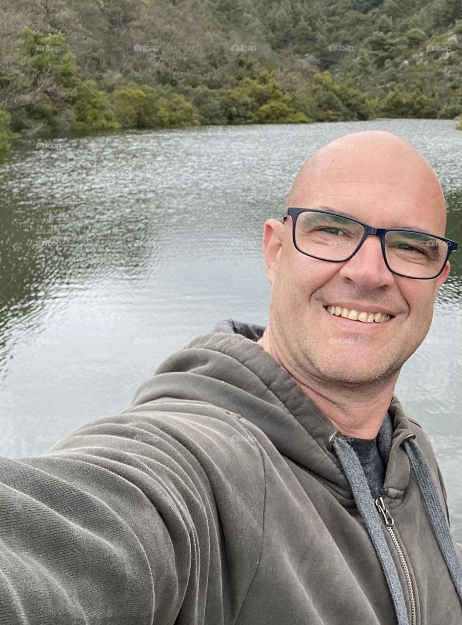 A bald headed man wearing glasses enjoying a walk in nature by a lake.