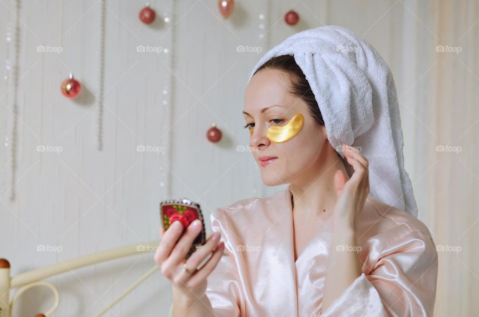 Young happy woman with a towel on head doing facial skin care procedures. Beauty routine. Cosmetic concept. Patches at the face.