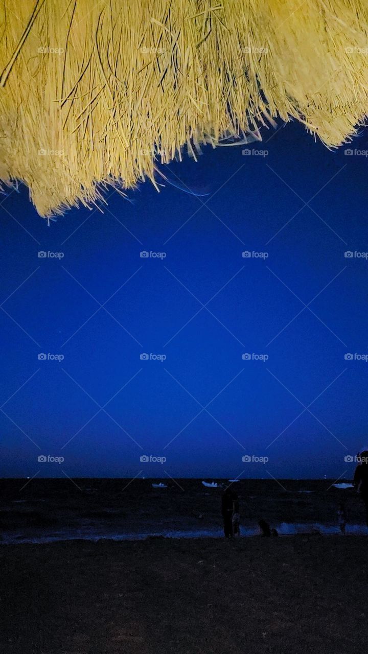 a serene nighttime beach scene. The top part of the image is dominated by a thatched umbrella, illuminated by a warm light, contrasting with the deep blue night sky. Below, the dark sea stretches across the horizon, with gentle waves visible as they
