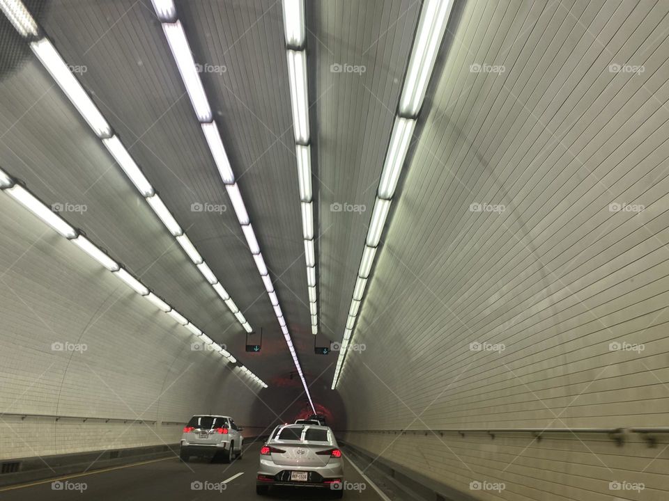 Two silver cars drive through an underground grey tunnel with fluorescent lights. 