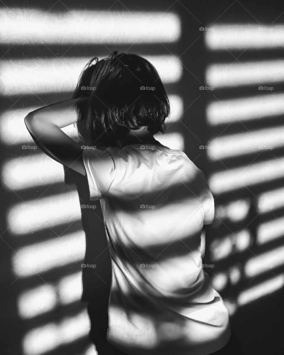 Black and white photograph of a girl standing against a wall with streaks of light and shadow from the blinds. The silhouette in a white T-shirt with her head down and her hand resting on the wall creates an atmosphere of thoughtfulness and lonelines