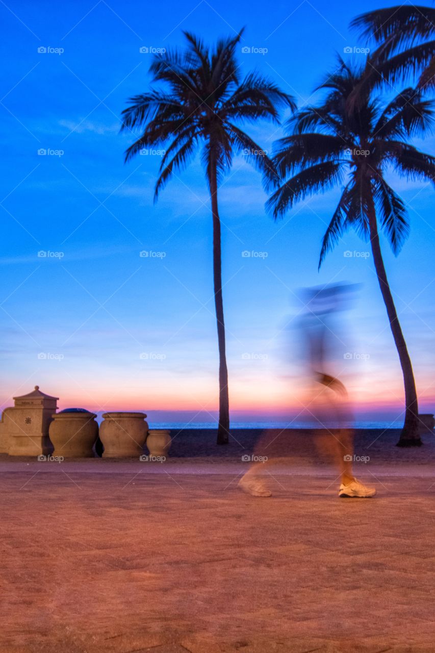 A walker is blurred as the sun creates silhouettes of the palm trees on an early morning sunrise at Hollywood Beach, Florida.  
