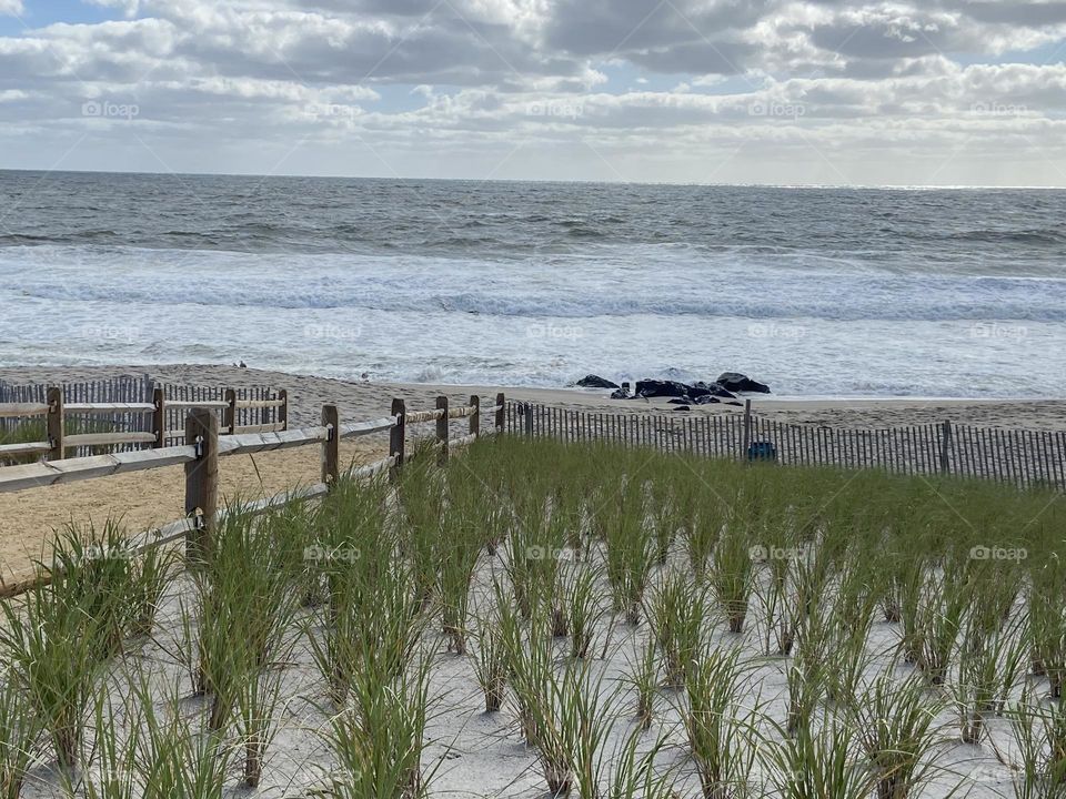 A view of the ocean and beach in Bay Head, NJ from an elevated platform with benches that leads to a sand path to the beach. Dune grass adds some green to the scene. Clouds fill the sky above the ocean on this September day.
