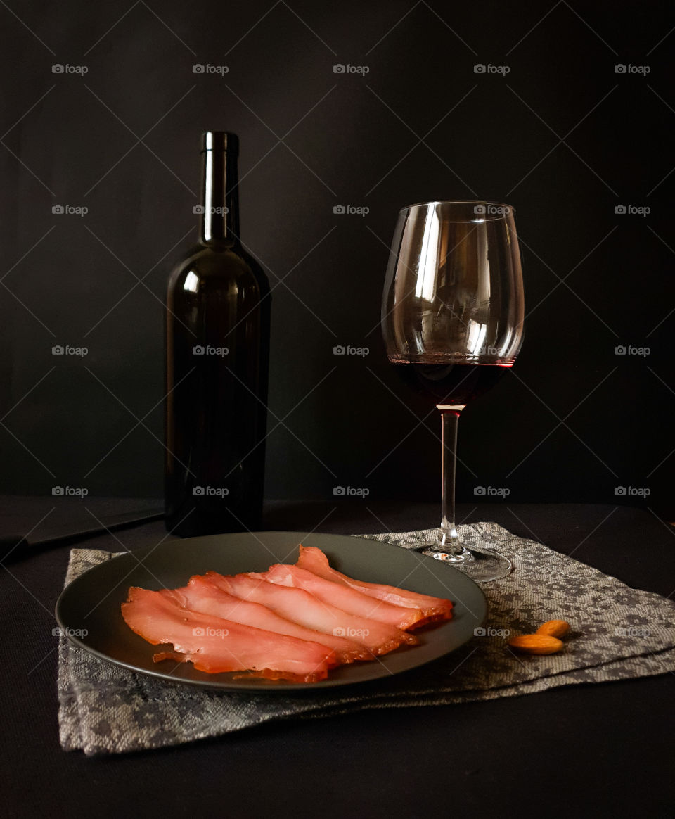 Dark still life. Black bottle of wine, a glass with red wine and a black plate with dried meat on a decorative ornamental napkin on a black background